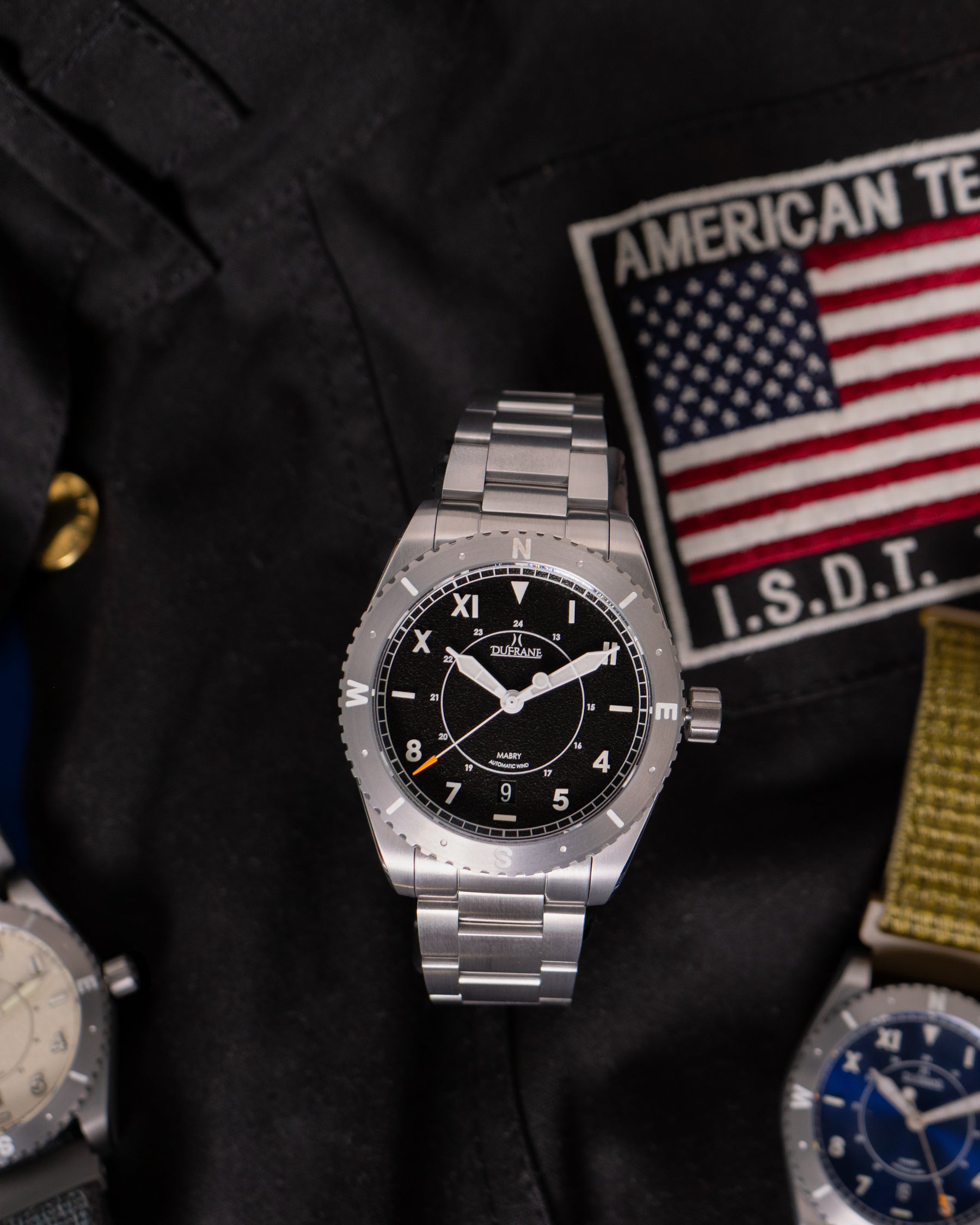 Silver watch with black dial on a dark surface with American flag and patches.